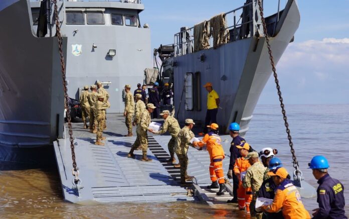 En un esfuerzo mancomunado entre la Armada de Colombia y la Gobernación de La Guajira, en articulación con el Programa Mundial de Alimentos (PMA), el Ejército Nacional, la Policía Nacional, la Cruz Roja Colombiana, la Defensa Civil, el ICBF, entre otras organizaciones, inició hoy la entrega de ayudas en el departamento de La Guajira, afectado por la segunda temporada de lluvias que azota al país en los últimos días. A primeras horas de la mañana emplayó en el sector conocido como “Amas Sai”, en Riohacha, el Buque de Desembarco Anfibio ARC “Golfo de Urabá” para iniciar el embarque de las ayudas que fueron dispuestas, con el fin de brindar asistencia humanitaria a las comunidades afectadas en la Alta Guajira, las cuales permanecen incomunicadas debido a las graves inundaciones de la región. A bordo del ARC “Golfo de Urabá” y de los camiones de Infantería de Marina de la Armada Nacional, que llegarán a siete puntos nodales de la Alta Guajira enfrentando condiciones adversas de tiempo y terreno, se transportarán más de 150 toneladas de elementos de primera necesidad y víveres, entre los que se encuentran alimentos, kits de aseo, agua potable, entre otros. Jairo Aguilar Deluque, gobernador de La Guajira, destacó la importancia de esta operación, señalando que “la unión de esfuerzos entre las instituciones y organizaciones permite atender de manera oportuna a quienes más lo necesitan, llevando esperanza a las comunidades afectadas”. En Riohacha fueron entregadas 2.7 toneladas de arroz gestionadas por la Armada de Colombia ante la Corporación “Minuto de Dios”, para iniciar la atención a los afectados de este municipio. Durante 15 días, el ARC “Golfo de Urabá” y los vehículos del Batallón de Movilidad de Infantería de Marina tendrán la misión de transportar y entregar ayudas humanitarias en Bahía Honda, Bahía Hondita, Castilletes, Karasúa, Puerto Bolívar, Puerto Estrella y Puerto López, con apoyo de 71 integrantes de las diferentes autoridades y entidades. Cabe resaltar que la Institución Naval desde la declaratoria de emergencia realizada por el Gobierno Nacional el pasado domingo 10 de noviembre, se encuentra desplegada con sus capacidades de empleo dual y su equipo humano para brindar apoyo humanitario y asistencia a la autoridad civil, en las comunidades del Caribe que han sido afectadas por este fenómeno. La Armada de Colombia, a través de la Fuerza Naval del Caribe, reitera su compromiso de contribuir a la consolidación territorial y al mejoramiento de la calidad de vida de los habitantes de la región, trabajando de manera articulada con entidades públicas y privadas.